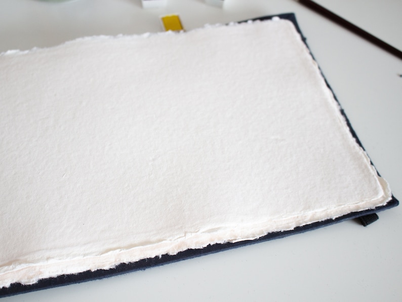 Puede incluir: Una hoja de papel blanco texturizado, en blanco, encuadernada en una cubierta azul oscuro. El papel tiene un borde irregular.