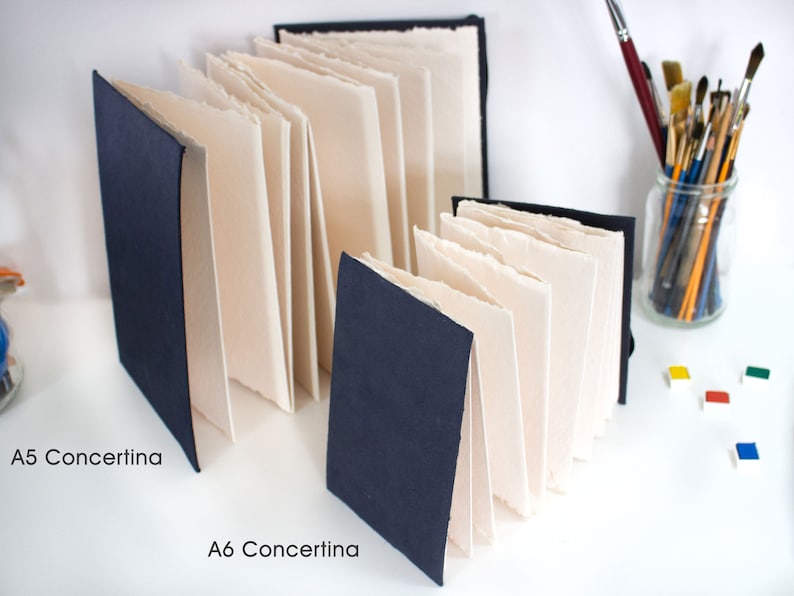 Puede incluir: Dos cuadernos concertina, uno A5 y otro A6, con cubiertas de color azul oscuro y p&aacute;ginas de papel blanco. Los cuadernos est&aacute;n sobre una superficie blanca con un frasco de pinceles y botes de pintura en el fondo.