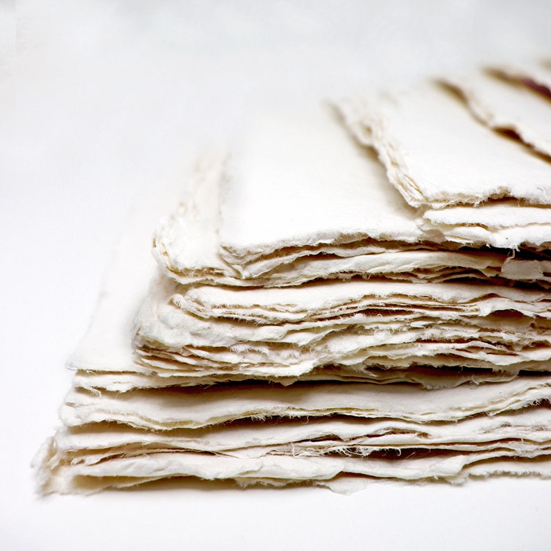 K&ouml;nnte beinhalten: Ein Stapel handgemachtes Papier mit einer rauen, strukturierten Oberfl&auml;che. Das Papier ist cremewei&szlig; und hat eine leicht unregelm&auml;&szlig;ige Form.