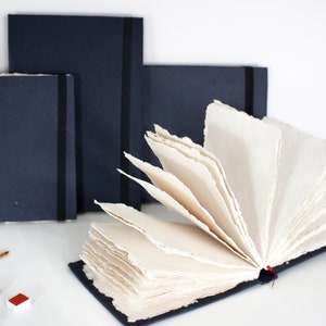 Puede incluir: Tres cuadernos negros con un marcador de cinta roja. El cuaderno de la derecha est&aacute; abierto, revelando papel hecho a mano de color crema con bordes irregulares.