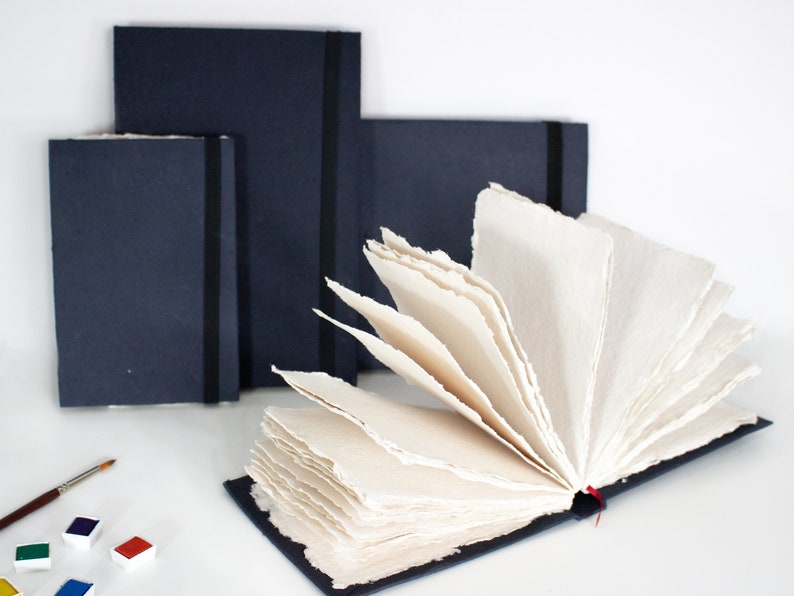 Puede incluir: Tres cuadernos negros con un marcador de cinta roja. El cuaderno de la derecha est&aacute; abierto, revelando papel hecho a mano de color crema con bordes irregulares.