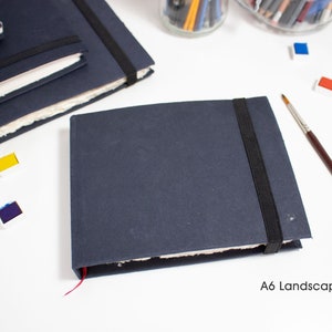 Puede incluir: Un cuaderno de dibujo azul oscuro con cierre el&aacute;stico negro y un marcador de cinta rojo. El cuaderno est&aacute; cerrado y sobre una superficie blanca. El texto "A6 Landscape" est&aacute; impreso debajo del cuaderno.
