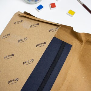 Puede incluir: Un cuaderno azul marino con cierre el&aacute;stico negro est&aacute; envuelto en papel marr&oacute;n con un patr&oacute;n repetido del texto "INDIGO ARTWAY".