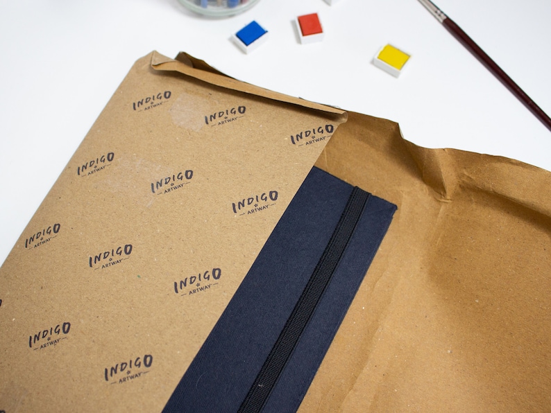 Puede incluir: Un cuaderno azul marino con cierre el&aacute;stico negro est&aacute; envuelto en papel marr&oacute;n con un patr&oacute;n repetido del texto "INDIGO ARTWAY".