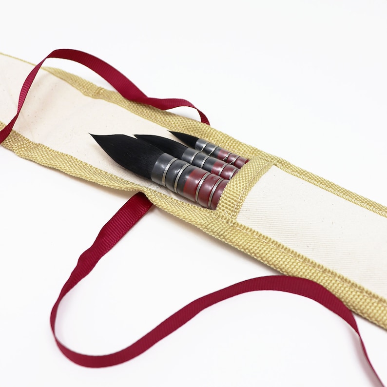 May include: A set of four black calligraphy brushes with red and silver handles are stored in a beige canvas case with a red ribbon.