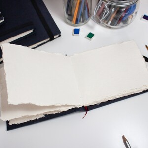Puede incluir: Un cuaderno de bocetos abierto y en blanco con una cubierta azul oscuro y un marcador de cinta roja. El cuaderno est&aacute; abierto sobre una superficie blanca con un frasco de l&aacute;pices, pinceles y acuarelas.