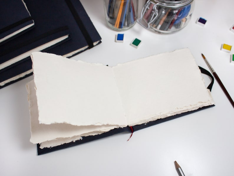 Puede incluir: Un cuaderno de bocetos abierto y en blanco con una cubierta azul oscuro y un marcador de cinta roja. El cuaderno est&aacute; abierto sobre una superficie blanca con un frasco de l&aacute;pices, pinceles y acuarelas.