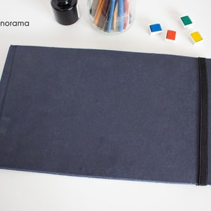 Puede incluir: Un cuaderno de dibujo azul oscuro con un cierre de banda el&aacute;stica negra. El cuaderno de dibujo est&aacute; acostado sobre una superficie blanca. La palabra "Panorama" es visible en la esquina superior izquierda de la imagen.