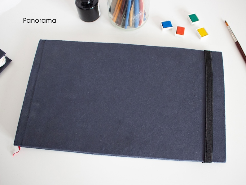 Puede incluir: Un cuaderno de dibujo azul oscuro con un cierre de banda el&aacute;stica negra. El cuaderno de dibujo est&aacute; acostado sobre una superficie blanca. La palabra "Panorama" es visible en la esquina superior izquierda de la imagen.