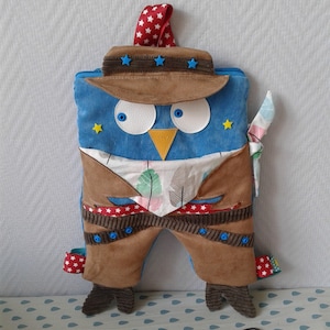 Può includere: Un giocattolo di peluche a forma di gufo blu e marrone che indossa un cappello da cowboy marrone con stelle blu. Il gufo ha un becco giallo e occhi bianchi. Il giocattolo è seduto su una superficie con motivo blu e bianco.