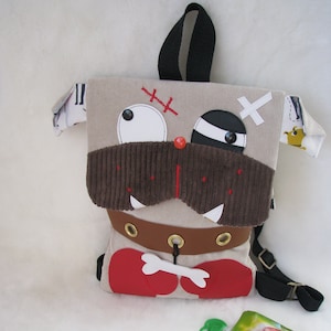 Puede incluir: Una mochila beige, marrón y roja con forma de perro con una correa negra. El perro tiene un ojo blanco, un parche negro en el ojo y una nariz de pana marrón. La mochila tiene un tirador de cremallera en forma de hueso y un cinturón marrón con ojales.