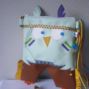Peut inclure: Un sac en forme de hibou bleu clair avec des accents marrons et une bordure en franges. Le sac a une sangle jaune, bleue et violette et comprend un collier de perles avec un couteau en bois et un fourreau en cuir.