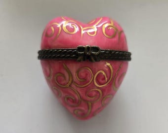 May include: A pink heart-shaped trinket box with a gold swirl design and a brown ribbon.