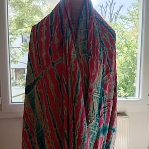 Peut inclure: Écharpe tie-dye vibrante aux tons de rouge, turquoise et vert-jaune. Le tissu est drapé sur un mannequin, mettant en valeur le design fluide. Le motif de l'écharpe présente des rayures verticales, créant un textile visuellement dynamique.