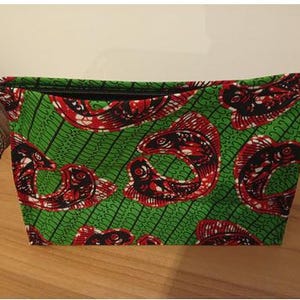 Op de afbeelding: Een rechthoekige groene make-up tas met een zwarte rits en een klein handvat. De tas is bedrukt met rode en zwarte visontwerpen op een groene achtergrond. De tas staat op een houten oppervlak.