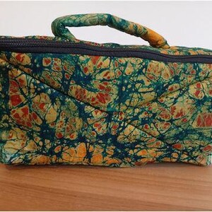 Op de afbeelding: Een rechthoekige, gewatteerde tas met een batikpatroon in tinten van teal, oranje en geel. De tas heeft een zwarte ritssluiting en een gewatteerde handgreep. De stof heeft een uniek, organisch ontwerp.