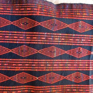 Puede incluir: Primer plano de un textil tejido con un patrón repetitivo. La tela presenta un fondo azul marino oscuro con bandas horizontales de rombos y líneas geométricas rojas y naranjas. La textura parece ligeramente áspera.