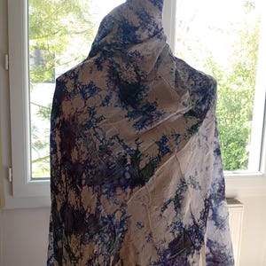 Peut inclure: Écharpe à motifs beige et bleu drapée sur un mannequin. L'écharpe présente un motif aquarelle avec des nuances de bleu, de violet et de vert. Le tissu semble léger et fluide, avec une texture froissée. L'écharpe est exposée près d'une fenêtre.