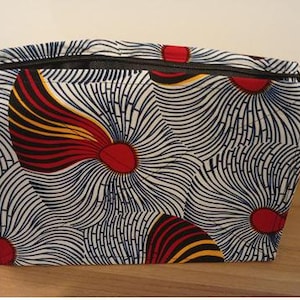 Op de afbeelding: Een rechthoekige make-uptas met een levendige Afrikaanse print. De tas heeft een zwarte ritssluiting en is versierd met een herhalend patroon van rode, gele en zwarte bloemmotieven op een witte en marineblauwe achtergrond.