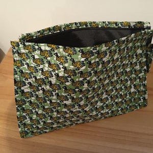 May include: A rectangular, open zipper pouch with a repeating pattern of yellow and black turtle-like shapes on a green and white background. The interior is black. The pouch is sitting on a light brown wooden surface.