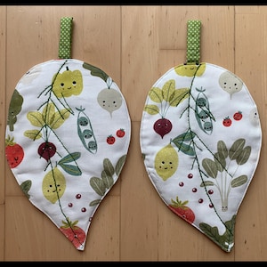 Peut inclure: Deux maniques en forme de feuille avec un fond blanc et un motif de légumes fantaisiste. Le motif comprend des citrons souriants, des radis, des pois et des tomates. Chaque manique a une boucle à pois verts pour l'accrochage.
