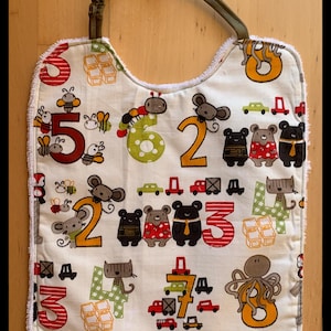 Puede incluir: Un babero para bebé de color crema con una variedad de ilustraciones coloridas. El babero presenta números, coches, abejas, ratones, osos y un pulpo. El babero tiene una correa verde y ribete blanco.