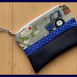 Peut inclure: Une petite pochette zippée bleue avec un tirette de fermeture éclair blanche. La pochette est faite d'un tissu bleu à pois blancs et d'un tissu gris avec un motif d'animaux de la ferme et un tracteur bleu. La pochette a un fond en cuir bleu.