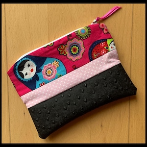 Puede incluir: Una bolsa de cremallera rosa con un diseño de tela negra, blanca y rosa. La bolsa tiene un fondo de tela negra con un patrón texturizado. La bolsa presenta una tela colorida con un patrón de muñecas rusas y flores.