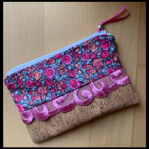 May include: A small pink and gray floral fabric pouch with a pink lace trim and a cork bottom.