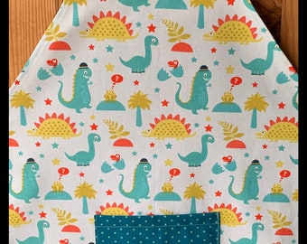 May include: A white apron with a teal blue polka dot pattern on the neck straps and pocket. The apron has a dinosaur print with green, yellow, and red dinosaurs on a white background.