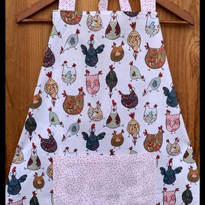 Può includere: Grembiule bianco con un motivo stravagante di galline multicolori. Il grembiule presenta un'ampia tasca e spalline con un design a pois rosa. Il grembiule è appeso a un'appendiabiti di legno.