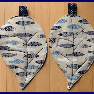 Può includere: Due presine a forma di foglia con un motivo a pesce. Le presine sono beige con un motivo a pesce blu navy e un anello blu navy a pois. I pesci sono in varie tonalità di blu e bianco.