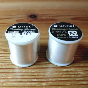 May include: Two spools of white beading thread. The spool on the left is labeled "MIYUKI Beading Thread 330dtex 100% NYLON Made in Japan LOT. BH1OR COL 1". The spool on the right is labeled "MIYUKI Beading Thread ビーズステッチ糸 50m (55yde.) フィルム:PET".