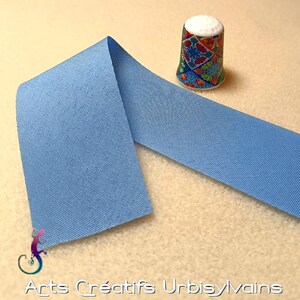 Peut inclure: Un ruban en tissu bleu clair plié en deux. Le ruban est texturé et a un léger brillant. Un dé à coudre coloré est visible en arrière-plan.