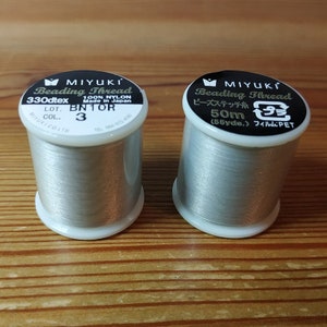 May include: Two spools of silver-colored beading thread. The spool on the left is labeled "MIYUKI Beading Thread 330dtex 100% NYLON Made in Japan LOT. BN1OR COL. 3". The spool on the right is labeled "MIYUKI Beading Thread ビーズステッチ糸 50m (55yde.) フィルム:PET".