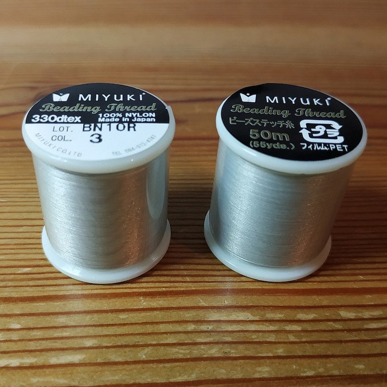 May include: Two spools of silver-colored beading thread. The spool on the left is labeled "MIYUKI Beading Thread 330dtex 100% NYLON Made in Japan LOT. BN1OR COL. 3". The spool on the right is labeled "MIYUKI Beading Thread ビーズステッチ糸 50m (55yde.) フィルム:PET".
