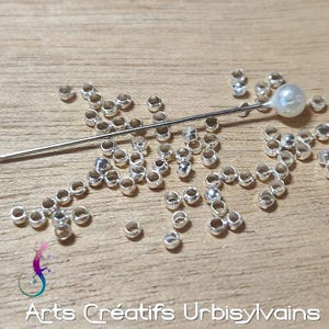 May include: A collection of small, silver-colored metal beads scattered on a wooden surface. A long, thin needle with a pearl-like head is also present. The image includes the text "Arts Créatifs Urbisylvains" at the bottom.