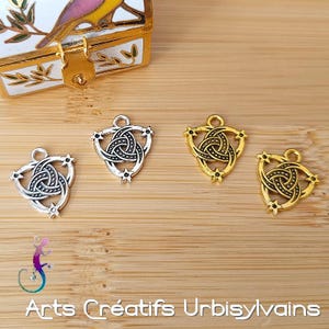 Puede incluir: Tres abalorios de plata y oro con un diseño de nudo celta negro. Los abalorios tienen forma de triángulo con tres puntas. Cada punta tiene un pequeño diseño de estrella.