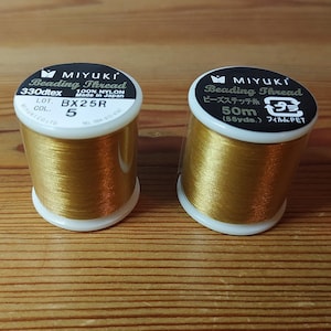 May include: Two spools of gold-colored beading thread. The spool on the left is labeled "MIYUKI Beading Thread 330dtex 100% NYLON Made in Japan LOT. BX25R 5 COL.". The spool on the right is labeled "MIYUKI Beading Thread ビーズステッチ糸 50m (55yds.) フィルム:PET"