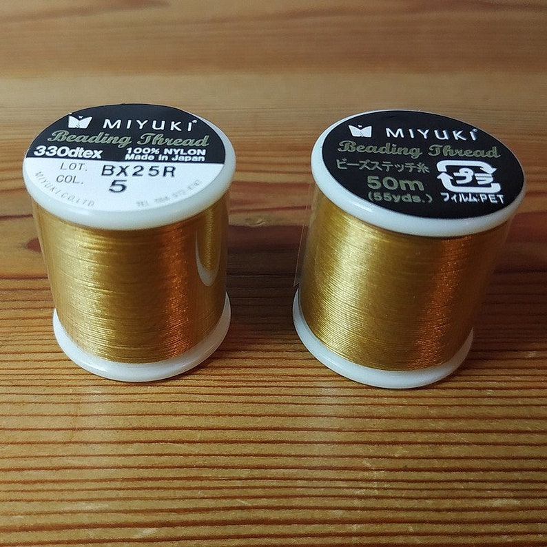 May include: Two spools of gold-colored beading thread. The spool on the left is labeled "MIYUKI Beading Thread 330dtex 100% NYLON Made in Japan LOT. BX25R 5 COL.". The spool on the right is labeled "MIYUKI Beading Thread ビーズステッチ糸 50m (55yds.) フィルム:PET"