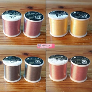 Pode incluir: Seis rolos de fio para miçangas Miyuki em várias cores, incluindo dourado, castanho, rosa e bege. Cada rolo é etiquetado com o nome da marca, o tipo de fio e o tamanho. Os rolos estão dispostos em duas filas de três.