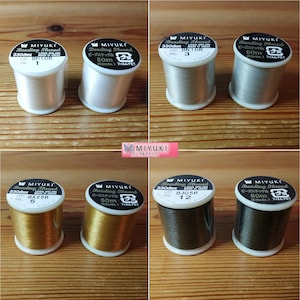 Puede incluir: Seis carretes de hilo para bordar Miyuki en varios colores, incluyendo blanco, plateado, dorado y negro. Cada carrete está etiquetado con el nombre de la marca, el tipo de hilo y el número de color. Los carretes están dispuestos en dos filas de tres.