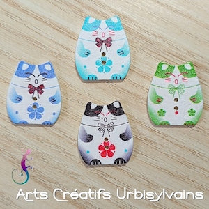 May include: Four wooden cat buttons with different coloured backgrounds. The buttons are painted with white faces, black eyes, and red bows. The backgrounds are blue, green, black and white, and light blue.