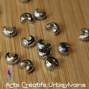 Peut inclure: Des perles à sertir en métal argenté, également appelées caches à sertir, sont éparpillées sur une surface en bois. Ces perles sont utilisées pour fixer les embouts de bijoux et créer une finition professionnelle.