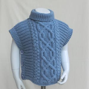 May include: A light blue knitted child's vest with a turtleneck collar and cable knit design. The vest has a button closure on the side and is displayed on a mannequin. The knitted vest is a cozy and stylish garment.