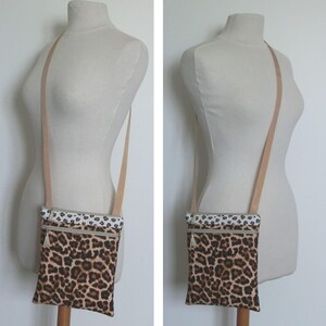 May include: A crossbody bag with a leopard print pattern. The bag has a beige strap and a white and black patterned top. It features two zippered compartments with silver-toned pulls. The bag is displayed on a mannequin.