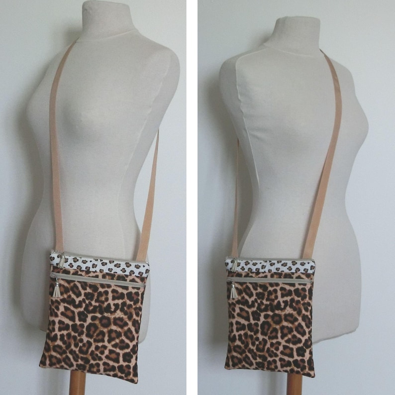 May include: A crossbody bag with a leopard print pattern. The bag has a beige strap and a white and black patterned top. It features two zippered compartments with silver-toned pulls. The bag is displayed on a mannequin.