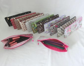 May include: A collection of rectangular fabric pencil cases featuring various patterns including hearts, florals, and geometric prints. Two open cases in the foreground reveal makeup brushes and colored pencils, each with a pink interior lining.