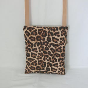 May include: A rectangular crossbody bag with a leopard print pattern in shades of brown and tan. The bag has a beige strap and a silver zipper with a tassel. The bag is designed to be worn over the shoulder.