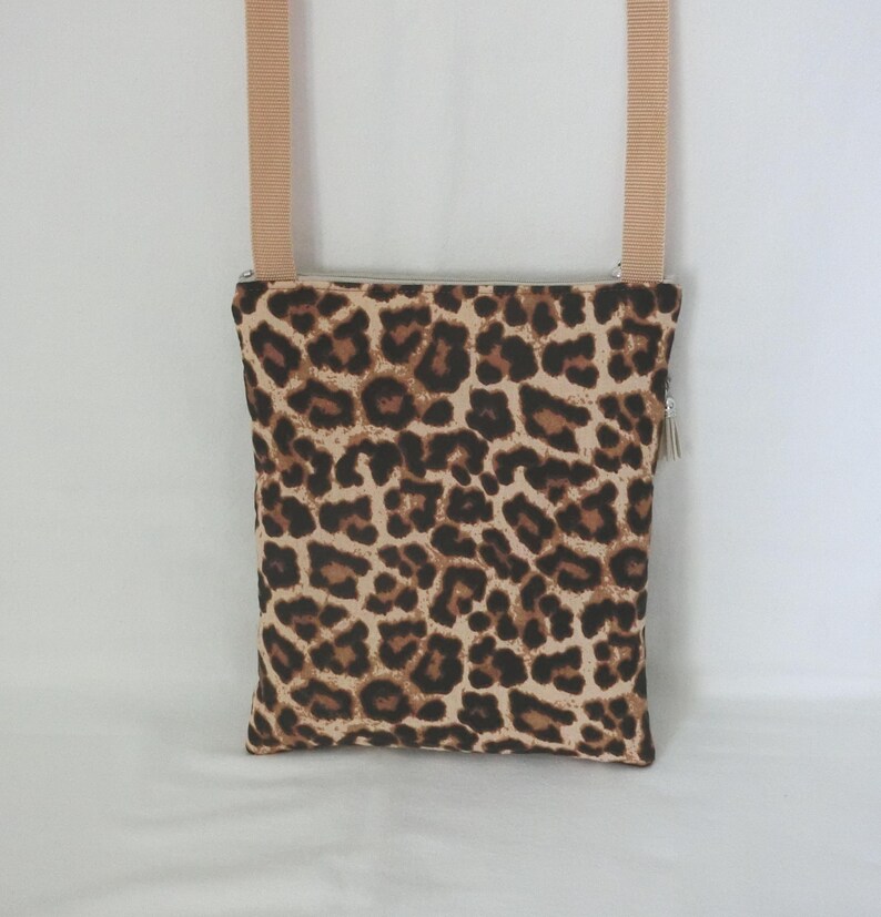 May include: A rectangular crossbody bag with a leopard print pattern in shades of brown and tan. The bag has a beige strap and a silver zipper with a tassel. The bag is designed to be worn over the shoulder.